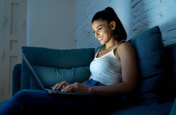 young attractive hispanic and relaxed woman at home smiling happy on sofa couch using laptop computer in dark evening light enjoying watching online movie or addicted to internet social network.