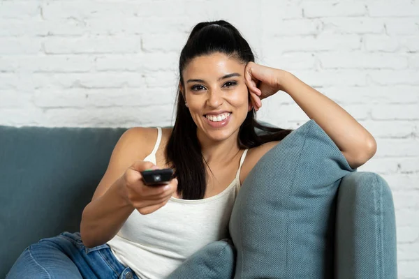 年轻美丽的西班牙妇女在牛仔裤在家独自看电视微笑坐在沙发沙发在客厅愉快和兴奋享受电视浪漫喜剧电影放松概念 — 图库照片