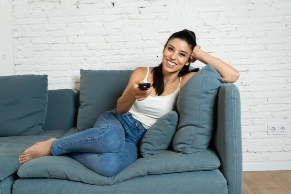 young beautiful Spanish woman in jeans home alone watching television smiling sitting on sofa couch in living room happy and excited enjoying a TV romantic comedy movie relaxed concept.