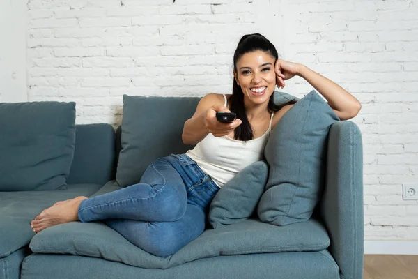 young beautiful Spanish woman in jeans home alone watching television smiling sitting on sofa couch in living room happy and excited enjoying a TV romantic comedy movie relaxed concept.