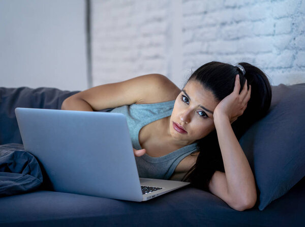 beautiful latin internet addicted woman working and surfing on her computer bored, sleepless and tired in her bed at home late at night in dark room looking surprised in addiction device concept.