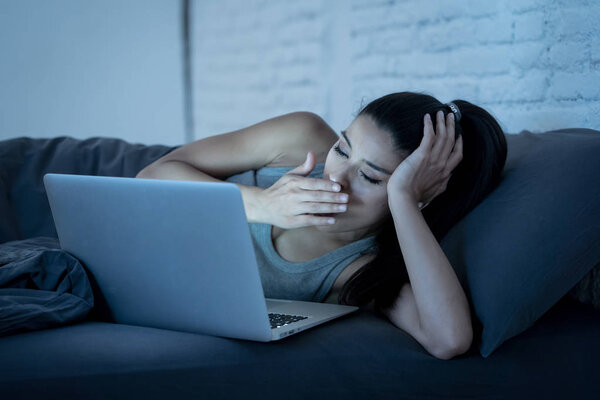 young beautiful hispanic internet addict woman in pajamas on bed at home bedroom working bored and tired with laptop computer late at night in dark room light yawning exhausted falling sleep.