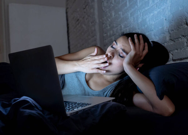 young beautiful hispanic internet addict woman in pajamas on bed at home bedroom working bored and tired with laptop computer late at night in dark room light yawning exhausted falling sleep.