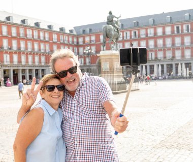 Beautiful happy senior couple taking photo with selfie stick in Plaza Mayor, Madrid, Spain. In retirement travel around Europe concept. clipart