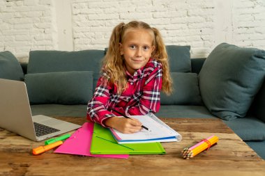 Sevimli beyaz neşeli İlköğretim öğrenci ödev yapmak ve evde oturma odasında dizüstü bilgisayar ile eğitim sırasında mutlu hissediyor