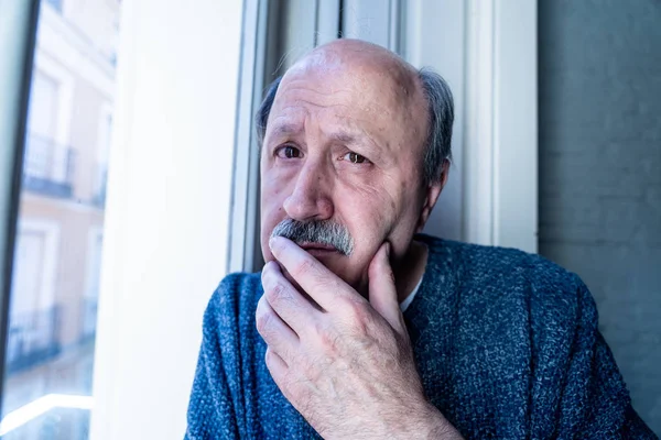 Old senior man looking throughout the window feeling confused and depressed alone at home in Aging Retirement Widower Dementia and Alzheimer concept.