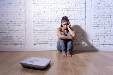 Young anorexic bulimic teenager woman sitting alone on ground looking at the scale worried and depressed in failing dieting and eating nutrition disorder concept. clipart
