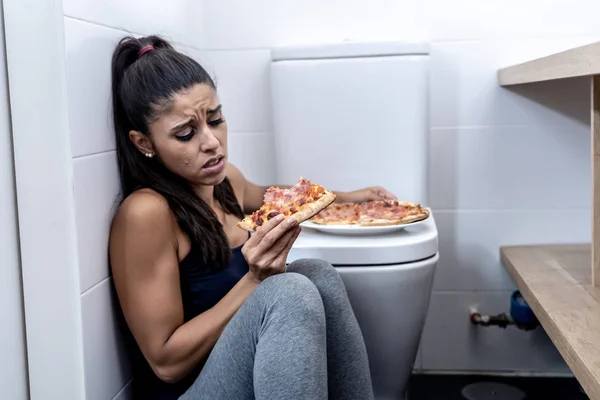 魅力的な若いと悲しい過食症若い女性有罪と摂食障害の拒食症と過食症の概念のトイレの横の床に座って食事が病気を感じ — ストック写真