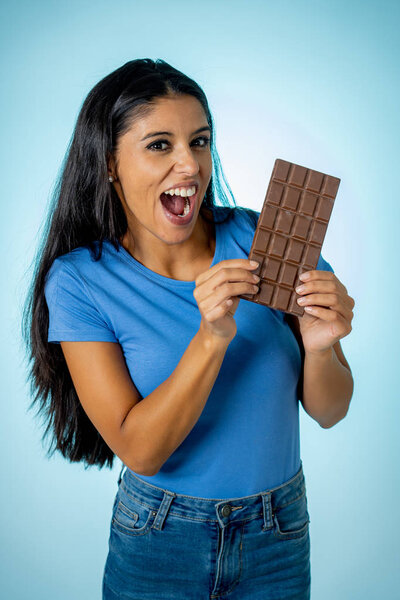 Beautiful young cute and happy latin woman in casual clothes holding big delicious chocolate tablet looking with temptation thinking if ignoring diet and eating sweet junk food isolated background