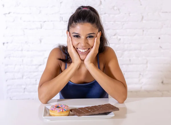 年轻迷人的拉丁妇女坐在桌子上吃巧克力和甜甜圈看起来兴奋和快乐在没有更多的饮食 糖和矮胖不健康的食物概念 — 图库照片