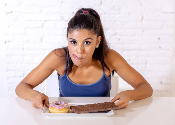 年轻迷人的拉丁妇女坐在桌子上吃巧克力和甜甜圈看起来兴奋和快乐在没有更多的饮食 糖和矮胖不健康的食物概念 — 图库照片