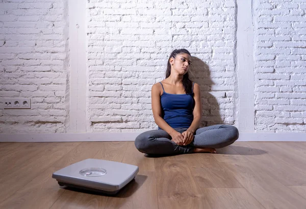 Young anorexic bulimic teenager woman sitting alone on ground looking at the scale worried and depressed in failing dieting and eating nutrition disorder concept.