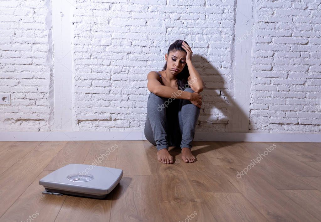 Young anorexic teenager woman sitting alone on ground looking at the scale worried and depressed in dieting and eating disorder concept