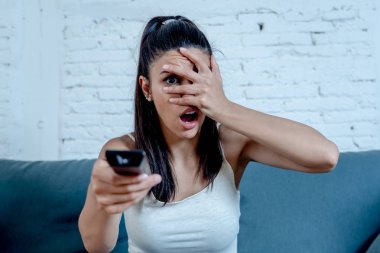 Güzel latin kadın korkmuş korkmuş ve yalnız evde oturma odasında kanepede oturup televizyon bir korku filmi izlerken tiksinti.