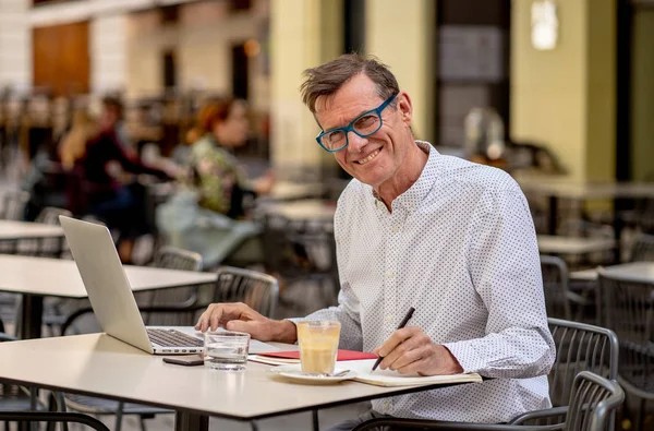 接続されてとどまる現代技術と起業家の創造的なビジネス概念を使用しての高齢者で屋外テラス コーヒー ショップ街にコーヒーを持ちながらコンピューターに取り組んで元気な笑顔の老人 — ストック写真