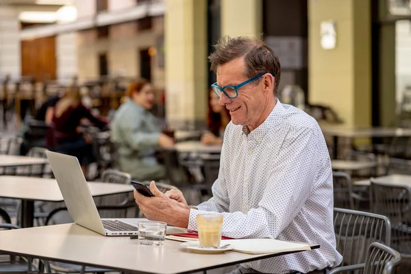 笑顔の老人が接続されてとどまる現代技術と起業家の創造的なビジネス概念を使用しての高齢者のテラス屋外コーヒー ショップでコンピューターで作業しながらスマート フォンをチェック — ストック写真