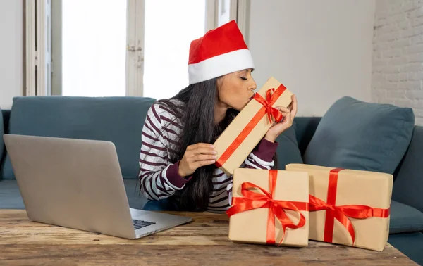 Woman wearing red sweater and santa claus hat choosing and buying christmas gifts on line using a laptop sitting on a couch in the living room at home excited about having all ready for christmas