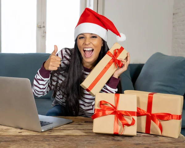 妇女穿着红色毛衣和圣诞老人帽子选择和购买圣诞礼物在线使用笔记本电脑坐在客厅里的沙发上兴奋着所有准备圣诞节 — 图库照片