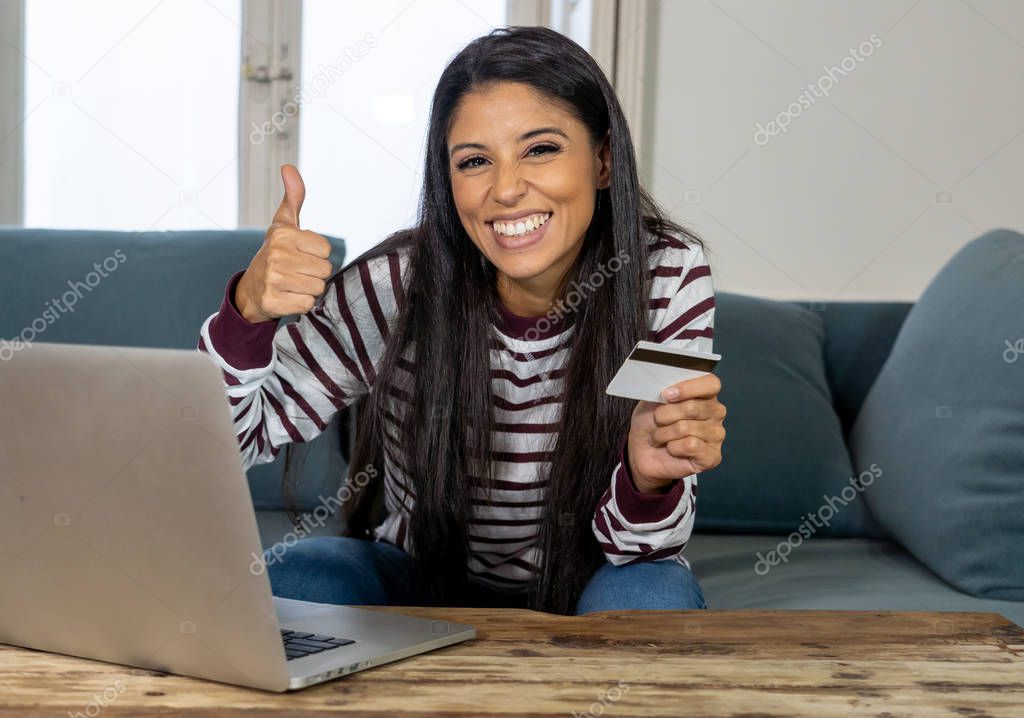 Attractive young latin woman holding credit card and using laptop computer buying on the internet at home in shopping on line business and technology digital marketing casual lifestyle concept.