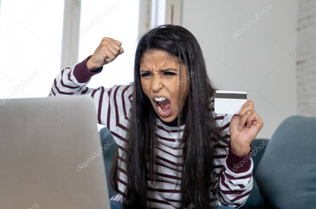Attractive young latin woman desperate and furious with credit card and laptop siting on sofa home in credit card problems debts online payment online shopping online banking bad financial situation.