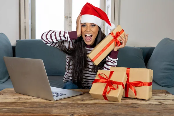 妇女穿着红色毛衣和圣诞老人帽子选择和购买圣诞礼物在线使用笔记本电脑坐在客厅里的沙发上兴奋着所有准备圣诞节 — 图库照片