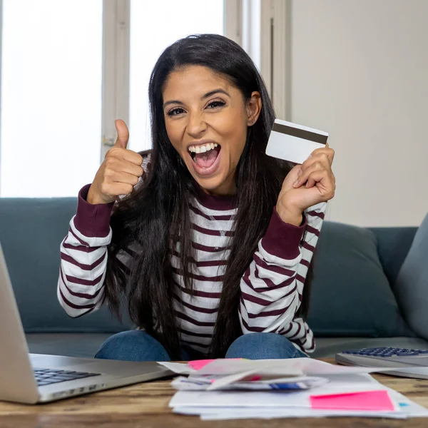 Cheerful attractive latin woman using credit card calculator and laptop paying bills at home looking relax in home finances paying bills online banking buying online and internet shopping.