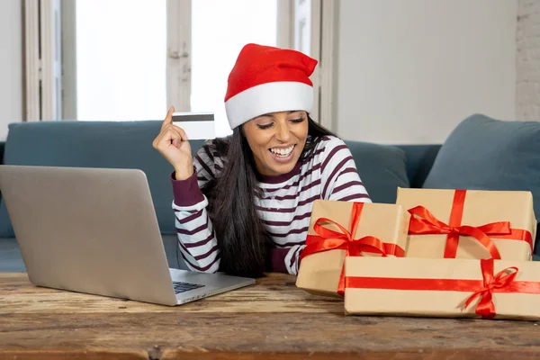 穿着红色毛衣和圣诞老人帽子持有信用卡选择和购买圣诞礼物使用笔记本电脑在家里兴奋与互联网销售和信用卡设施在线购物的妇女 — 图库照片