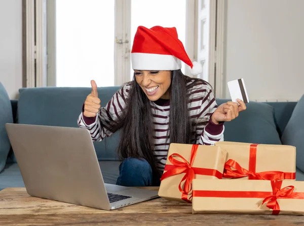 穿着红色毛衣和圣诞老人帽子持有信用卡选择和购买圣诞礼物使用笔记本电脑在家里兴奋与互联网销售和信用卡设施在线购物的妇女 — 图库照片