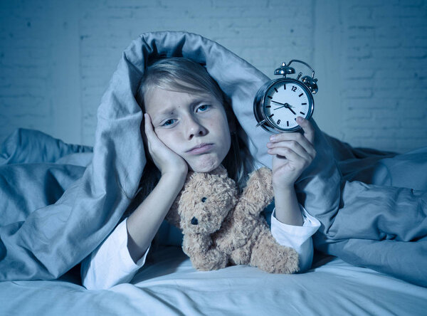 Симпатичная бессонная маленькая девочка лежала в постели, показывая будильник, выглядящий усталым, имеющий проблемы со сном, оставаясь спящим ночью или просыпаясь слишком рано в Бессонница Бессонница Бессонница и расстройства сна
.