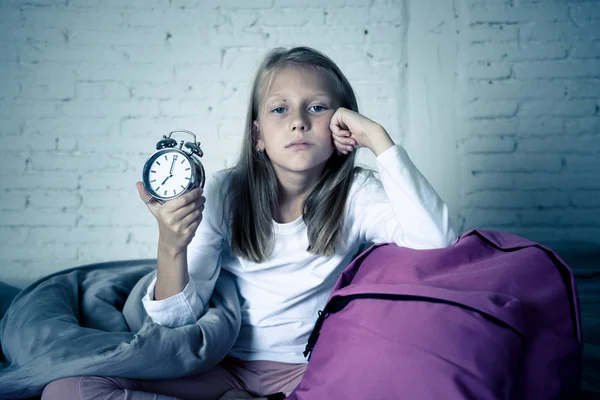 Beautiful blonde little girl sad sleepless and angry showing alarm clock time to get ready for school in difficulties waking up in the morning Children insomnia and sleeping disorders concept.