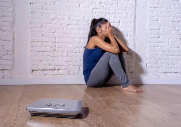 Young anorexic teenager woman sitting alone on ground looking at the scale worried and depressed in dieting and eating disorder concept