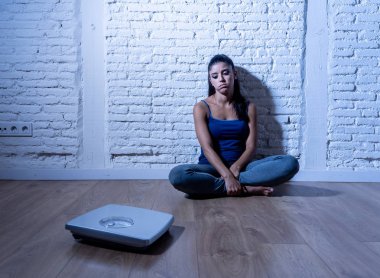 Young anorexic teenager woman sitting alone on ground looking at the scale worried and depressed in dieting and eating disorder concept clipart