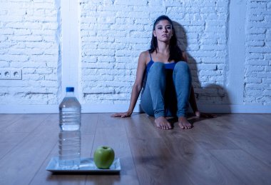 Desperate dieting young woman sitting on the ground looking sad at one apple fruit and water as symbol of crazy diet willpower and nutrition disorder. clipart