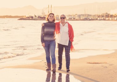 Mutlu kıdemli anne ve yetişkin kızı harcama zaman gülüyor ve gün batımı mutlu aile anları nesiller emeklilik ve insanlar kavramı ışığında kumsalda yürürken el ele birlikte portre.