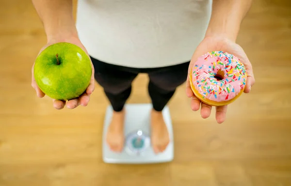 Close up of woman on scale holding on hands apple and doughnut making choice between healthy unhealthy food dessert while measuring body weight in Nutrition Health care Diet and temptation concept.