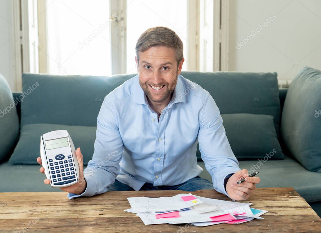 Young attractive caucasian business man accounting costs, charges, taxes and mortgage on sofa at home with credit card and calculator for paying bills looking happy and relaxed with business success