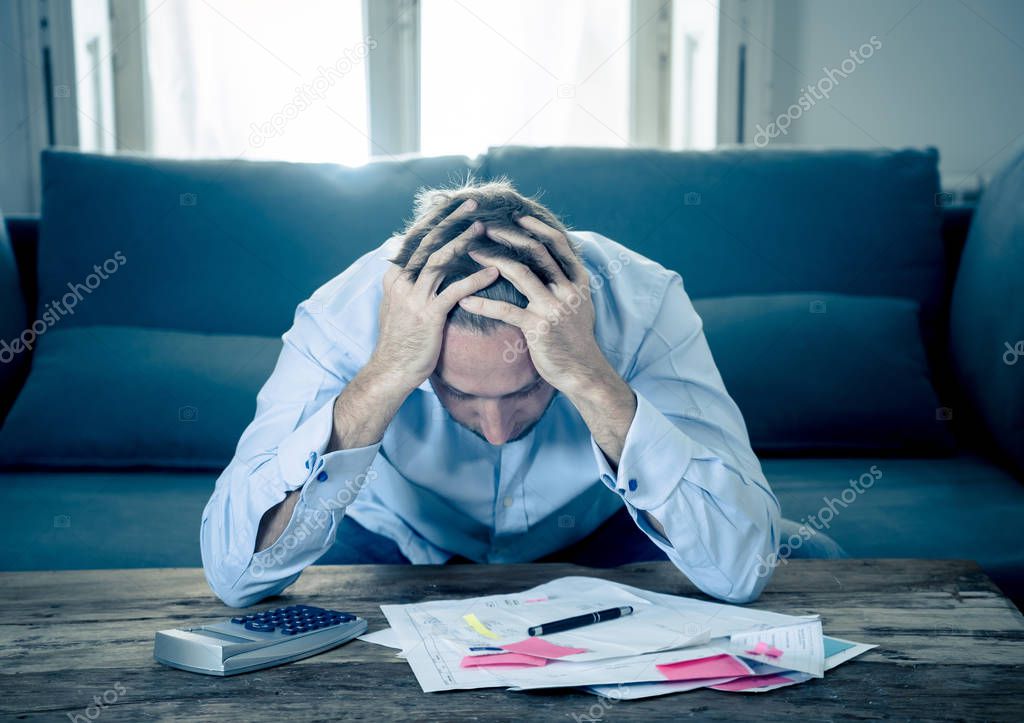 Worried and desperate entrepreneur young man calculating bills tax expenses and counting business or home finances sitting on couch at home in paying off debts banking and financial problems concept.