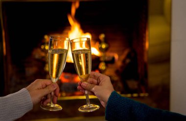 Kış tatili tatilinde ve sevgililer günü tatilinde ve yıldönümünde birlikte aşk mutlu anları kutlayan açık ahşap şömine tarafından kadeh kaldıran iki şampanya kadehi tutan romantik çift yakın.