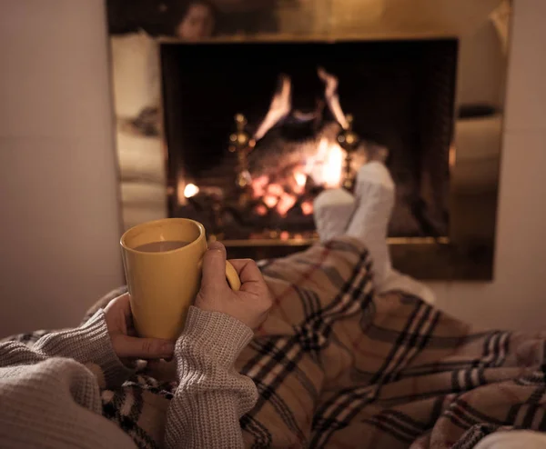 Close up image of woman sitting under the blanket by cozy fireplace warming up her feet in woolen socks relaxing with cup of hot chocolate drink at home in cold winter vacations in happy moments.