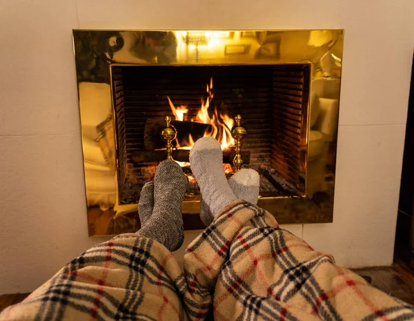 Close up image of couple sitting under the blanket by cozy fireplace warming up feet in woolen socks relaxing with cup of hot drink at home in cold winter vacations, happy moments and christmas.