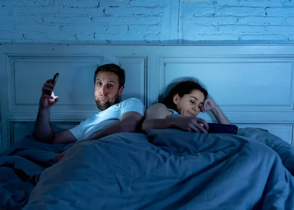 Sad man and woman married couple using their smart mobile phone in bed at night ignoring each other as strangers in relationship and communication problems and internet social network addiction.