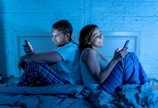 Couple in bed using phones, husband frustrated upset and unsatisfied while internet addict wife ignores him and enjoys mobile phone in social network addiction and relationship domestic problems.