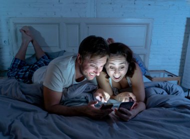 Smiling attractive young men and woman couple using smartphone lying on bed together. Happy boyfriend and girlfriend checking social networks or mobile app in gaming and technology addiction concept.
