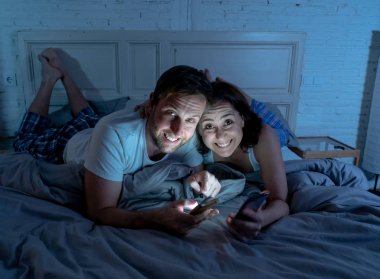 Smiling attractive young men and woman couple using smartphone lying on bed together. Happy boyfriend and girlfriend checking social networks or mobile app in gaming and technology addiction concept.