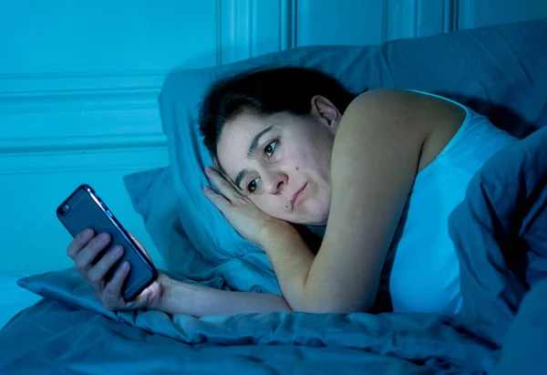 Internet Addicted young beautiful woman chatting and surfing on the internet using her smart phone sleepy bored and tired late at night in mobile addiction and insomnia concept