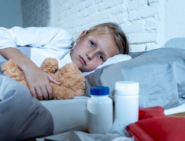 Sweet sick cute girl feeling sick lying in bed with medicines thermometer hot water bag suffering from Cold and Winter Flu Virus Sneezing Running Nose and Gripe Disease Symptoms in Child Health Care.