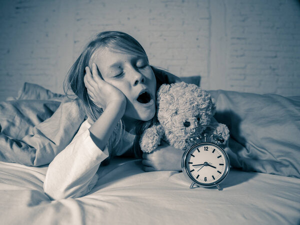 Симпатичная бессонная маленькая девочка лежала в постели, показывая будильник, выглядящий усталым, имеющий проблемы со сном, оставаясь спящим ночью или просыпаясь слишком рано в Бессонница Бессонница Бессонница и расстройства сна
.