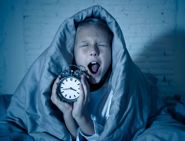 Симпатичная бессонная маленькая девочка лежала в постели, показывая будильник, выглядящий усталым, имеющий проблемы со сном, оставаясь спящим ночью или просыпаясь слишком рано в Бессонница Бессонница Бессонница и расстройства сна
.