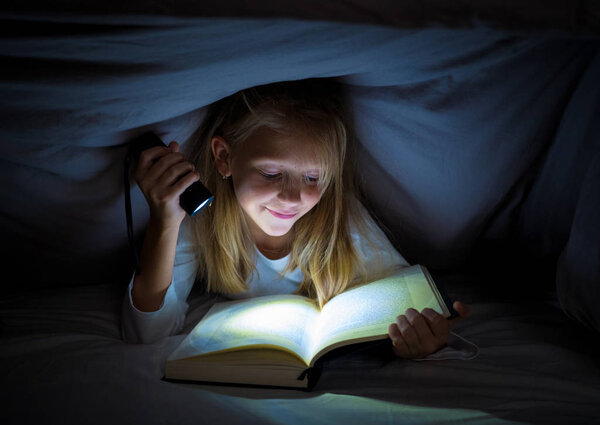 Happy smart girl reading a fairytale book lying in the dark under the cover bed holding a lantern in Reading skills Literature School Success and Education concept.