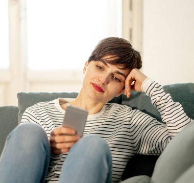Yeni teknoloji ve sosyal ağ kavramı kullanarak mutlu çekici kısa saçlı Genç kadın sohbet ve hatta, dating evde kanepede oturan sosyal medya uygulamaları kullanarak mobil akıllı telefon.